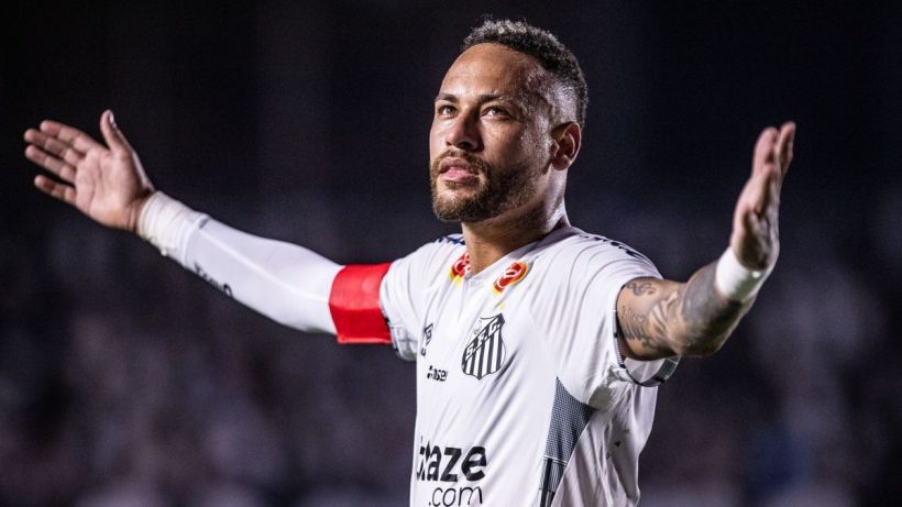 A soccer player in a white jersey with the Santos FC logo, raising his arms in celebration. He has a captain's armband and is in a dynamic pose on the field.