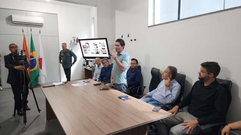 Reunião em uma sala com um grupo de pessoas, onde um homem está falando em pé ao lado de uma tela que exibe apresentações, enquanto outros homens escutam sentados à mesa.