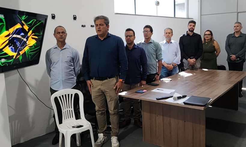 Grupo de pessoas em pé em uma sala, com um painel mostrando a bandeira do Brasil ao fundo. Eles estão de pé em frente a uma mesa de madeira, ouvindo o hino nacional.