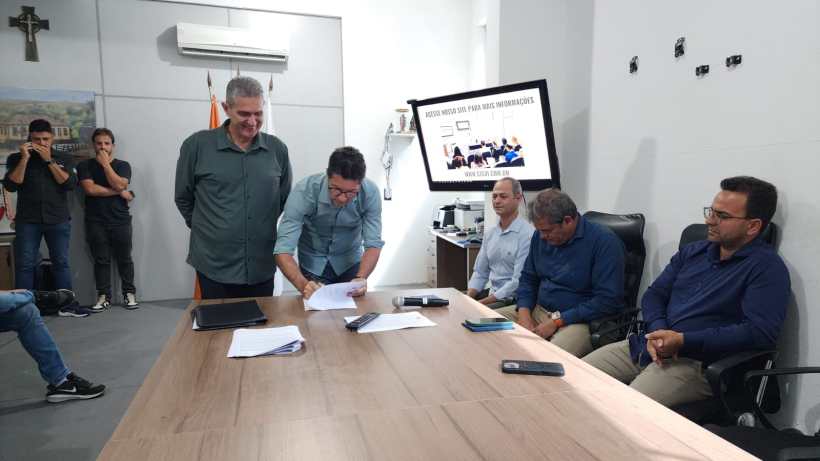 Reunião formal com várias pessoas ao redor de uma mesa, onde um homem está assinando documentos enquanto os outros observam.
