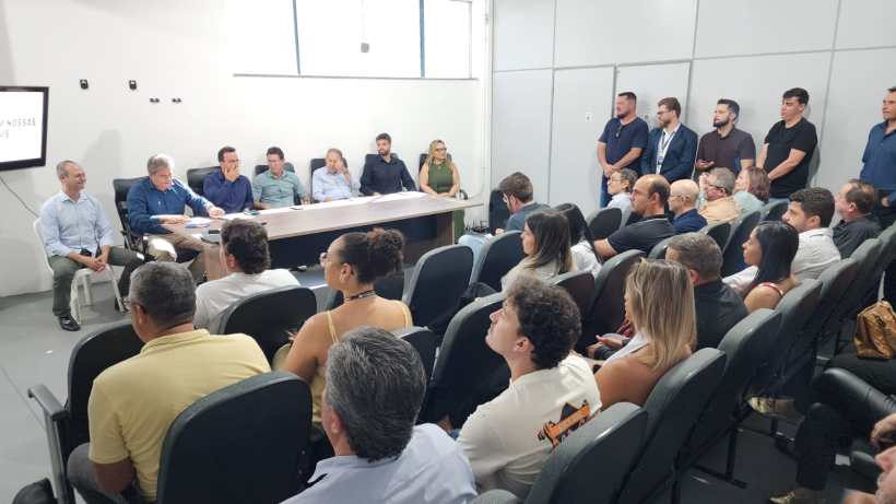 Reunião com várias pessoas sentadas e em pé, discutindo em uma sala com uma mesa na frente. Há um apresentador e um grupo de ouvintes, incluindo algumas mulheres.