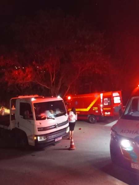 Cena noturna com veículos de emergência, incluindo uma ambulância e um caminhão de serviço, com luzes vermelhas piscando, em uma estrada arborizada.