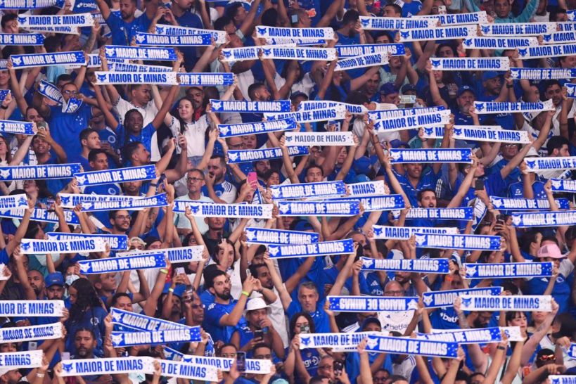 A large crowd of fans wearing blue clothing, holding up scarves that read 'MAIOR DE MINAS' in support of their team.
