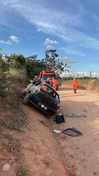 Carro acidentado em estrada de terra, inclinado em um barranco, com equipes de resgate e um caminhão de bombeiros ao fundo.