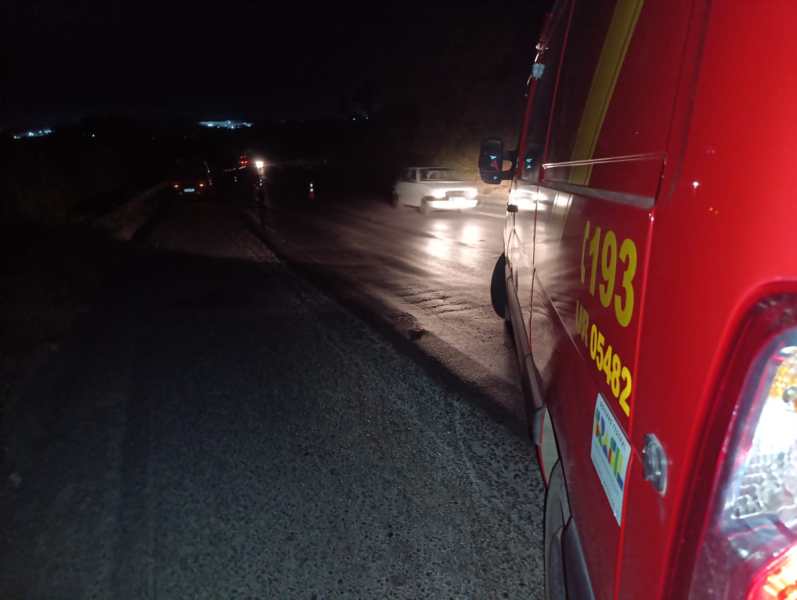 Veículo de emergência vermelho estacionado à beira da estrada à noite, com luzes de outros carros visíveis ao fundo.