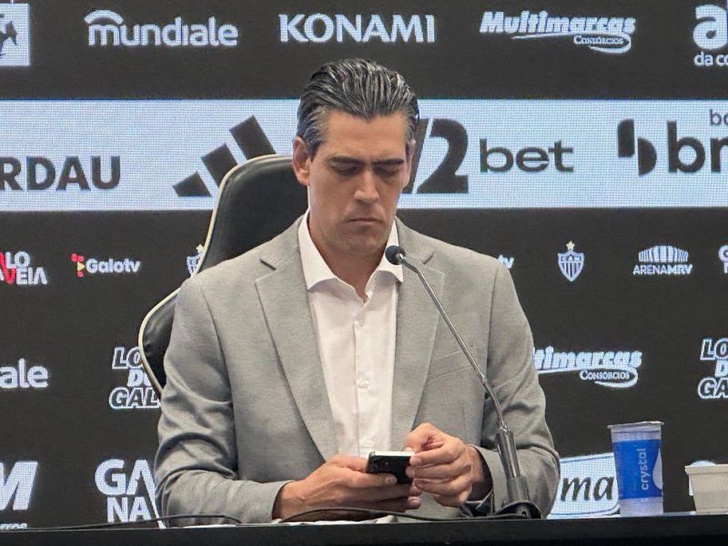 A person in a gray blazer sits at a press conference table, looking down at a smartphone with a serious expression. A microphone is positioned in front of them, and various logos are visible in the background.