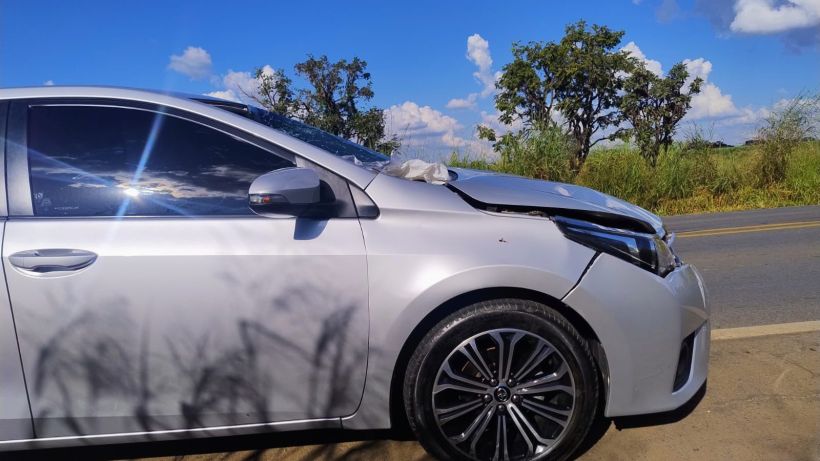 Um carro prateado danificado estacionado à beira da estrada, com uma paisagem verde e céu azul ao fundo.