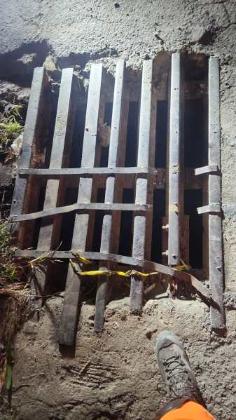 Uma grade de ferro sobre um buraco na calçada, com folhas secas ao redor e uma bota visível na parte inferior da imagem.