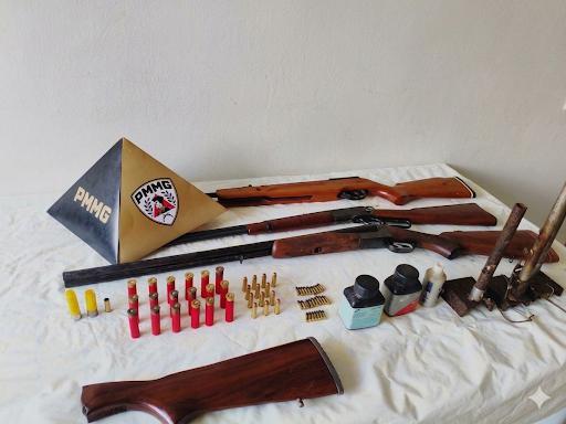 Exibição de armas, munições e materiais relacionados dispostos sobre uma superfície branca, incluindo um triângulo com a sigla PMMG.