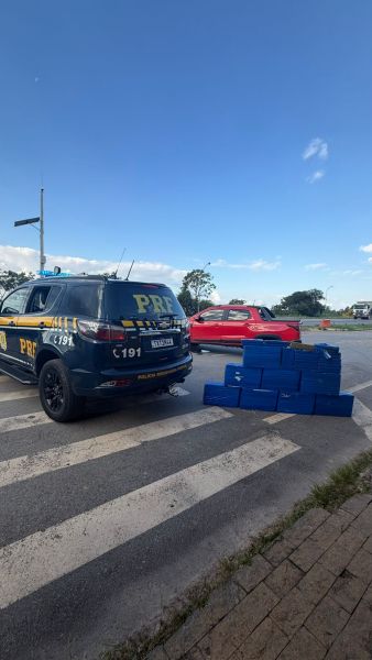 Veículo da PRF parado em uma estrada, com caixas azuis empilhadas na faixa de pedestres e um carro vermelho ao fundo.
