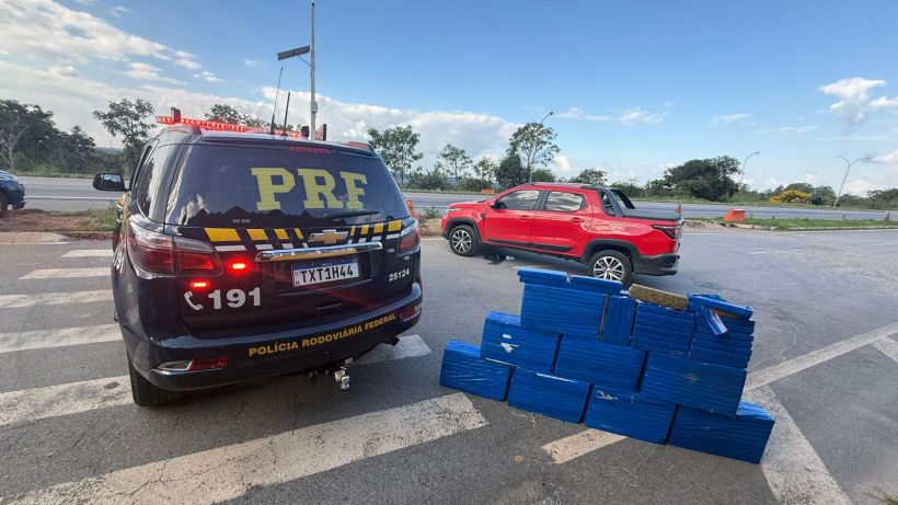 Veículo da Polícia Rodoviária Federal (PRF) estacionado ao lado de um carro vermelho, com fardos azuis empilhados no chão.