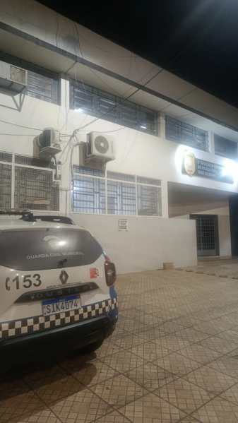 Viatura da polícia estacionada em frente a uma delegacia à noite, com ar-condicionados visíveis nas janelas.