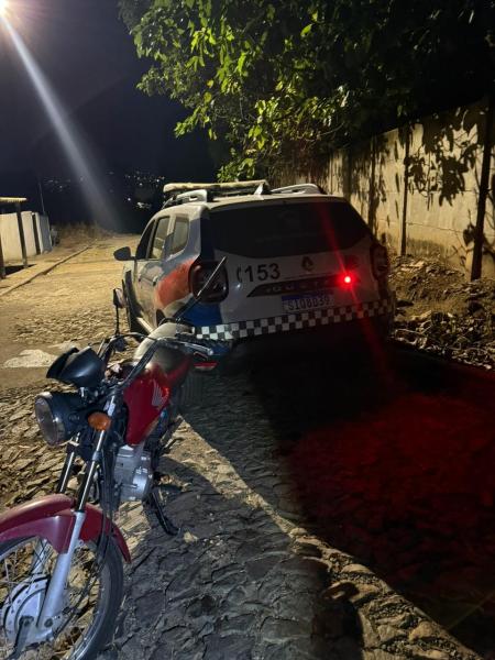 Veículo de polícia estacionado à noite em uma rua de paralelepípedos, ao lado de uma moto vermelha.