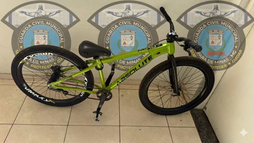 Bicicleta verde da marca Absolute, modelo N1, com pneus pretos, exposta em um ambiente interno com emblemas da Guarda Civil Municipal ao fundo.