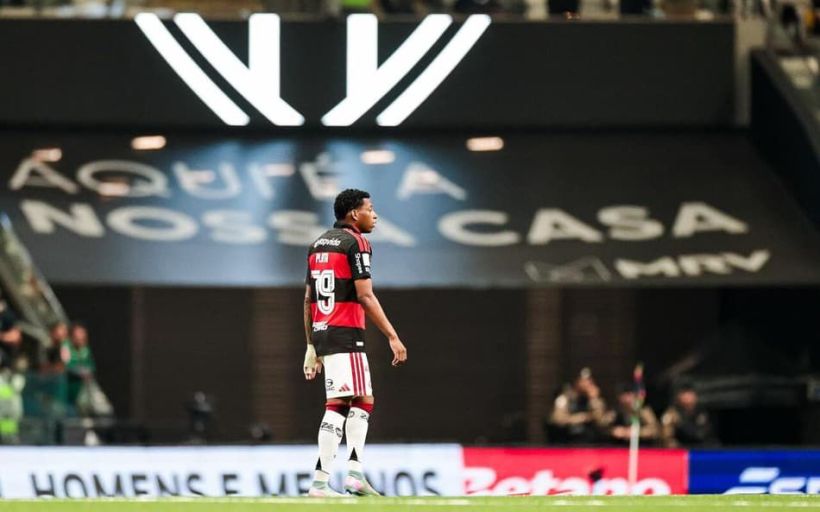 Jogador do time Flamengo em campo, com uniforme rubro-negro, olhando para um grande banner que diz 'Aqui é a nossa casa'.