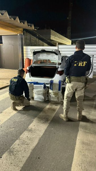 Dois agentes da PRF inspecionando o porta-malas de um carro em horário noturno, com pacotes suspeitos visíveis.