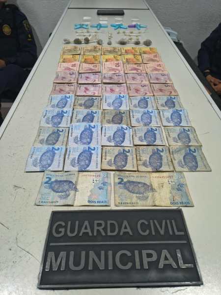 Notas de real organizadas em uma mesa, com destaque para uma placa da Guarda Civil Municipal ao fundo.