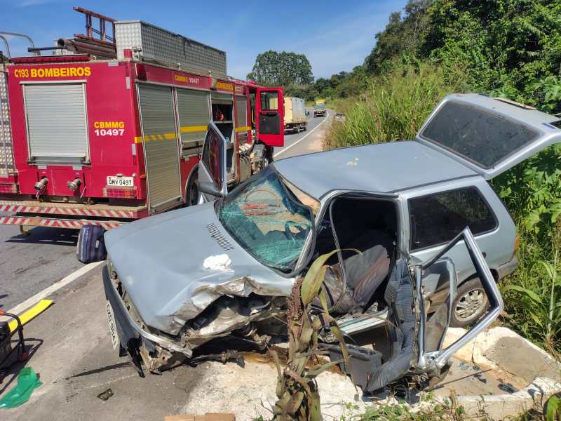 Carro acidentado na beira da estrada, com a parte frontal danificada e portas abertas, ao lado de um caminhão de bombeiros estacionamento.