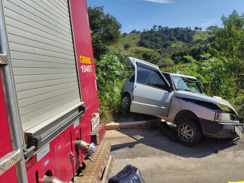 Carro acidentado ao lado de uma estrada, com capô danificado e portas abertas. Em primeiro plano, parte de um caminhão de bombeiros é visível.