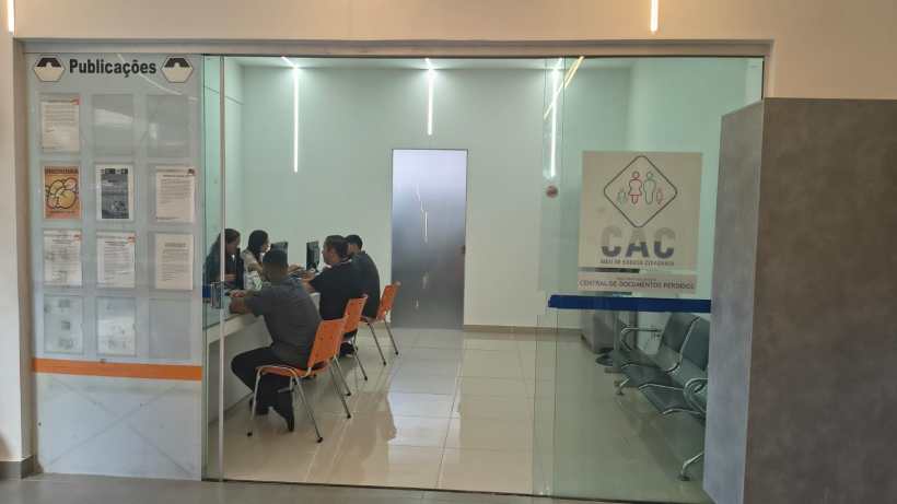 Sala de atendimento com diversas pessoas sentadas à mesa, em um ambiente iluminado e moderno, com uma placa indicando 'SAC' na entrada.