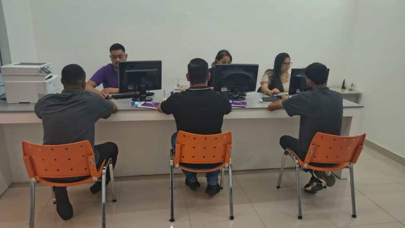 Pessoas atendendo em um balcão de atendimento, com quatro clientes sentados em cadeiras laranjas e empregados trabalhando em computadores.