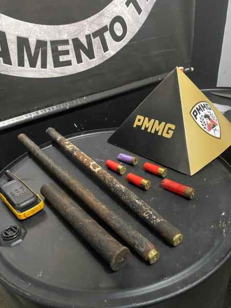 Conjunto de tubos de metal, cartuchos coloridos e um objeto em forma de pirâmide com o logo PMMG sobre uma superfície preta.