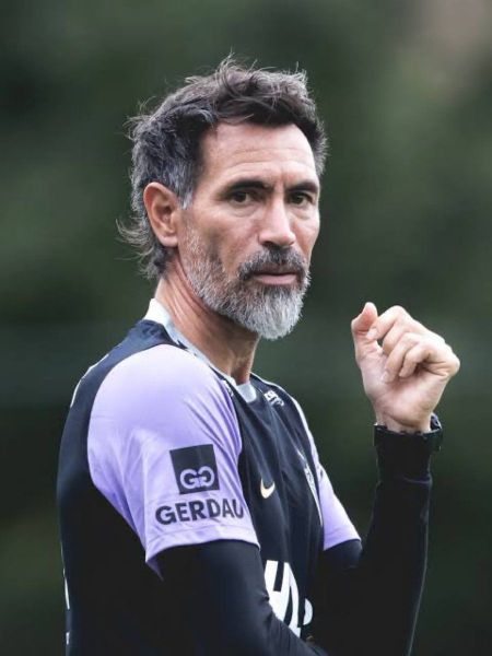 Homem com cabelos grisalhos, usando uniforme de treinamento de futebol, olhando para a câmera com expressão séria.