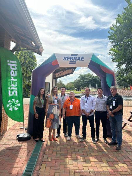 Grupo de pessoas posando em frente a um arco com a marca Sebrae, em um evento ao ar livre, com bandeira da Sicredi ao lado.