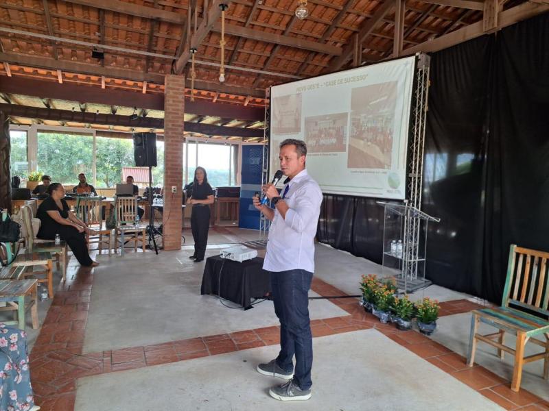 Homem apresentando em evento, com um microfone na mão, ao lado de uma mulher e uma apresentação em tela. O ambiente é um salão com mesas e cadeiras, decorado com plantas.