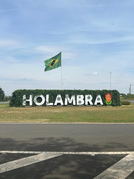 Placa de boas-vindas em Holambra com letras brancas, cercada por vegetação, e a bandeira do Brasil ao fundo.