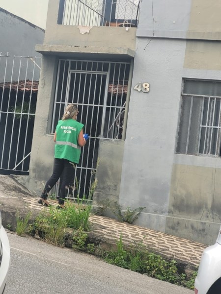 Mulher vestindo colete verde com a inscrição 'Assistência Social' caminhando em direção a um portão em frente a uma casa numerada 48.