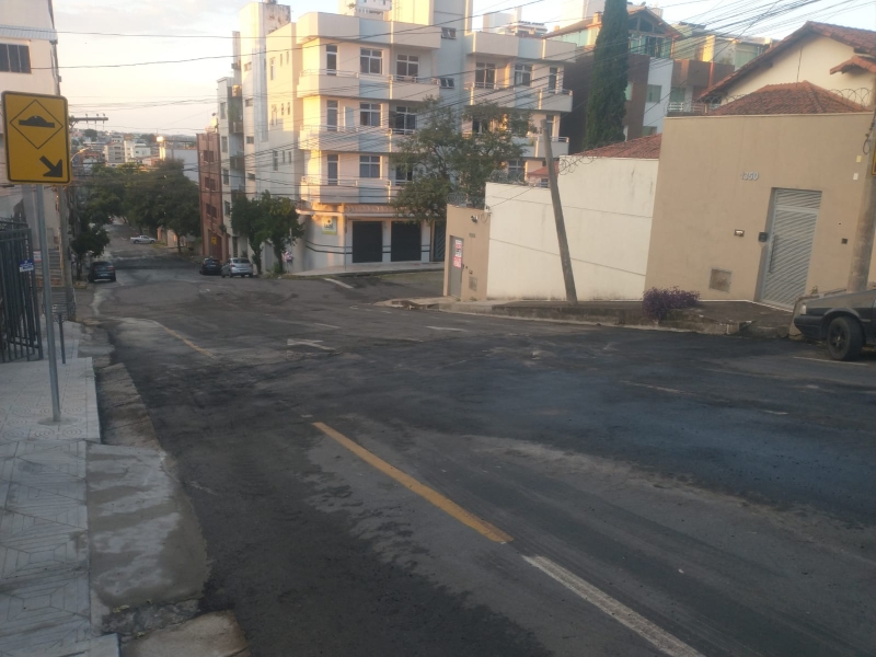 Vista de uma rua em uma área urbana, mostrando edifícios residenciais e carros estacionados, além de sinalização de tráfego e pavimentação irregular.