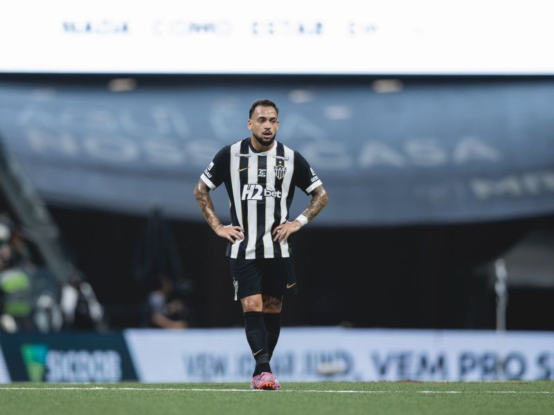 A soccer player in a black and white striped jersey stands on the field, appearing contemplative during a match.
