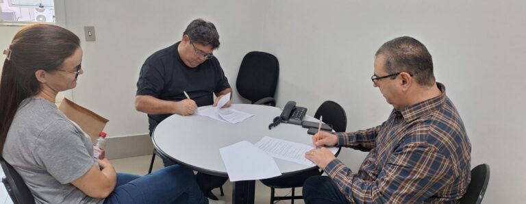 Três pessoas em uma reunião, sentadas em torno de uma mesa redonda, com papéis e canetas, em um ambiente interno iluminado.