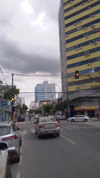 Rua com semáforo amarelo, carros circulando e prédios ao fundo em um dia nublado.