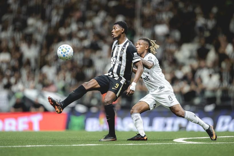 A soccer match scene featuring two players; one in a black and white striped jersey attempting to control the ball, while the other in a white jersey is closely defending.