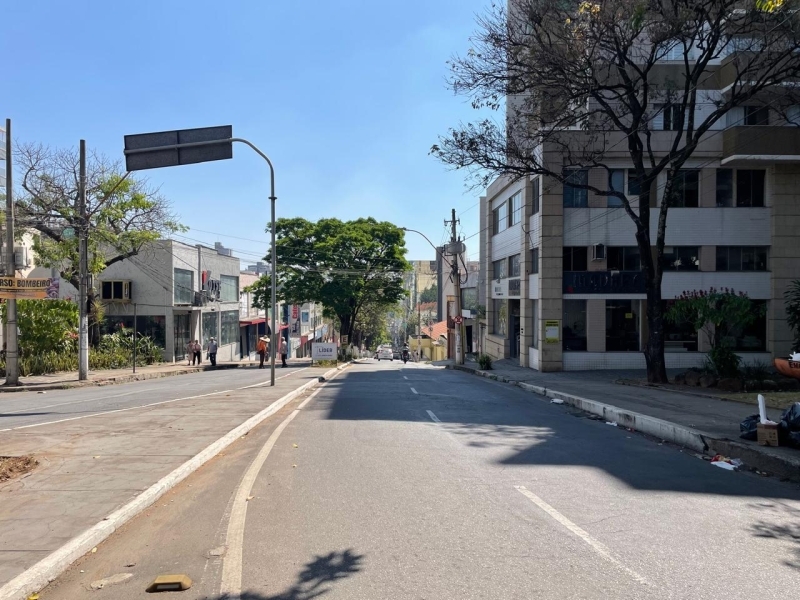 Rua urbana com prédios de ambos os lados, árvores e placas de sinalização sob céu azul claro.
