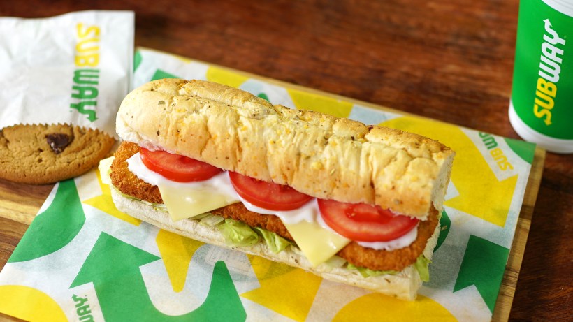 A Subway sandwich with chicken patties, lettuce, tomato, mayonnaise, and cheese on a toasted hoagie roll, accompanied by a chocolate chip cookie and a Subway drink cup on a wooden cutting board.