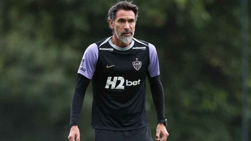 A middle-aged man with a beard, wearing a black athletic shirt with purple sleeves and a logo, stands on a soccer field with a focused expression.