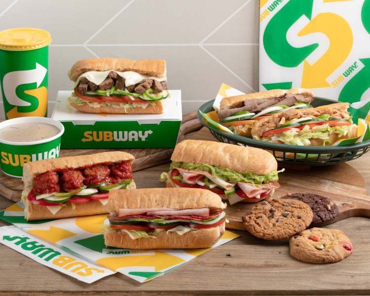 A display of various Subway sandwiches, including meatball, turkey, and veggie options, alongside a cup of drink and cookies on a wooden surface.