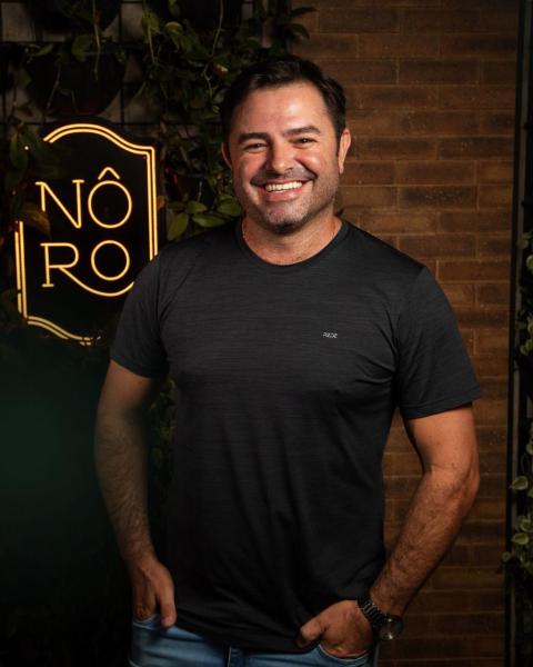 Homem sorrindo em frente a um fundo de tijolos e uma placa iluminada com o texto 'NÔ RO'.