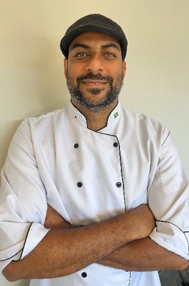 Chef sorrindo com braços cruzados, usando um uniforme branco de cozinheiro e um chapéu escuro.