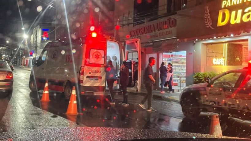 Ambulância e viatura da polícia paradas em uma rua à noite, com cones de sinalização e pessoas se movendo nas proximidades.
