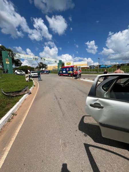Imagem de uma estrada com um veículo branco à frente e uma ambulância azul em movimento. Ao fundo, há uma entrada de local com várias pessoas e nuvens no céu.