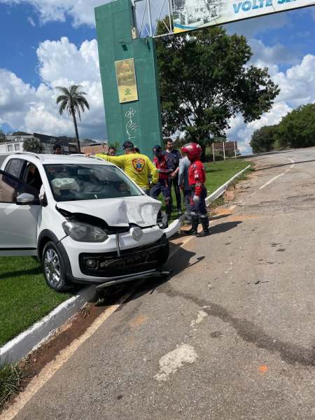Um carro branco batido está estacionado em uma calçada, com danos visíveis na frente. Várias pessoas estão ao redor do veículo, algumas usando capacetes, sugerindo que pode ter ocorrido um acidente de motocicleta.