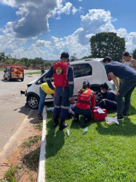 Equipe de resgate prestando atendimento a um acidente de trânsito ao lado de um carro branco, com pessoas ao redor ajudando.