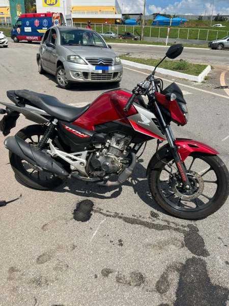Moto Honda em destaque, modelo naked, com design moderno e cor vermelha. Ao fundo, um carro estacionado em uma pista urbana.