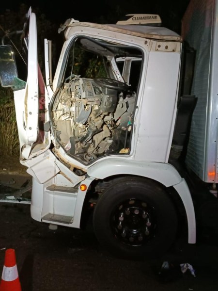 Caminhão acidentado com a cabine danificada, exposta e algumas peças visíveis, em uma estrada à noite.