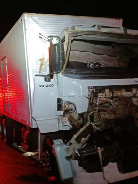 Caminhão danificado após um acidente, com a parte frontal severamente destruída e luzes de emergência ao fundo.