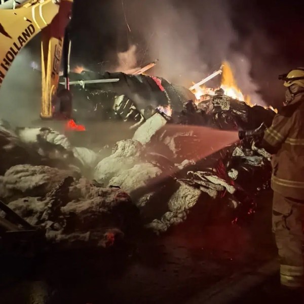 Bombeiros combatendo incêndio com mangueira, enquanto uma escavadeira move os destroços queimados.